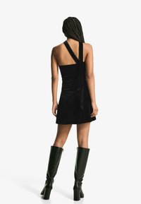 Žena s opletenými vlasmi, odetá v čiernych jednoramenných mini šatách a čiernych čižmách na vysokých podpätkoch po kolená, stojí s chrbtom k objektívu na bielom pozadí.