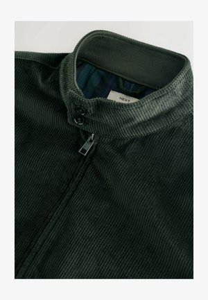 Veste en velours côtelé vert foncé avec un col haut, deux boutons et une fermeture éclair. L'intérieur présente une doublure à carreaux contrastante.