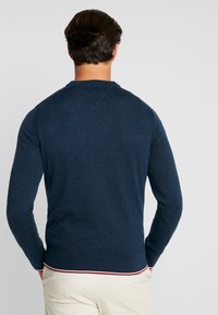 Jersey azul marino con textura, mangas largas, puños de canalé y bajo a rayas rojas y blancas en contraste. Vista trasera mostrada.