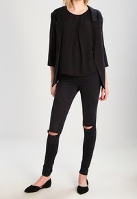 Pantaloni neri aderenti con strappi al ginocchio, abbinati a una blusa nera ampia e a un giacca nera strutturata. Completato con scarpe basse nere.