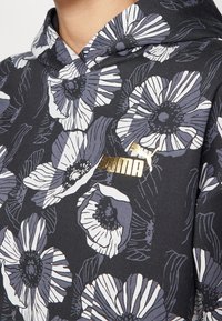 Schwarze Kapuzenjacke mit großem weißen und grauen Blumenmuster und kleinem goldenen Puma-Logo auf der Brust, getragen von einer Person.