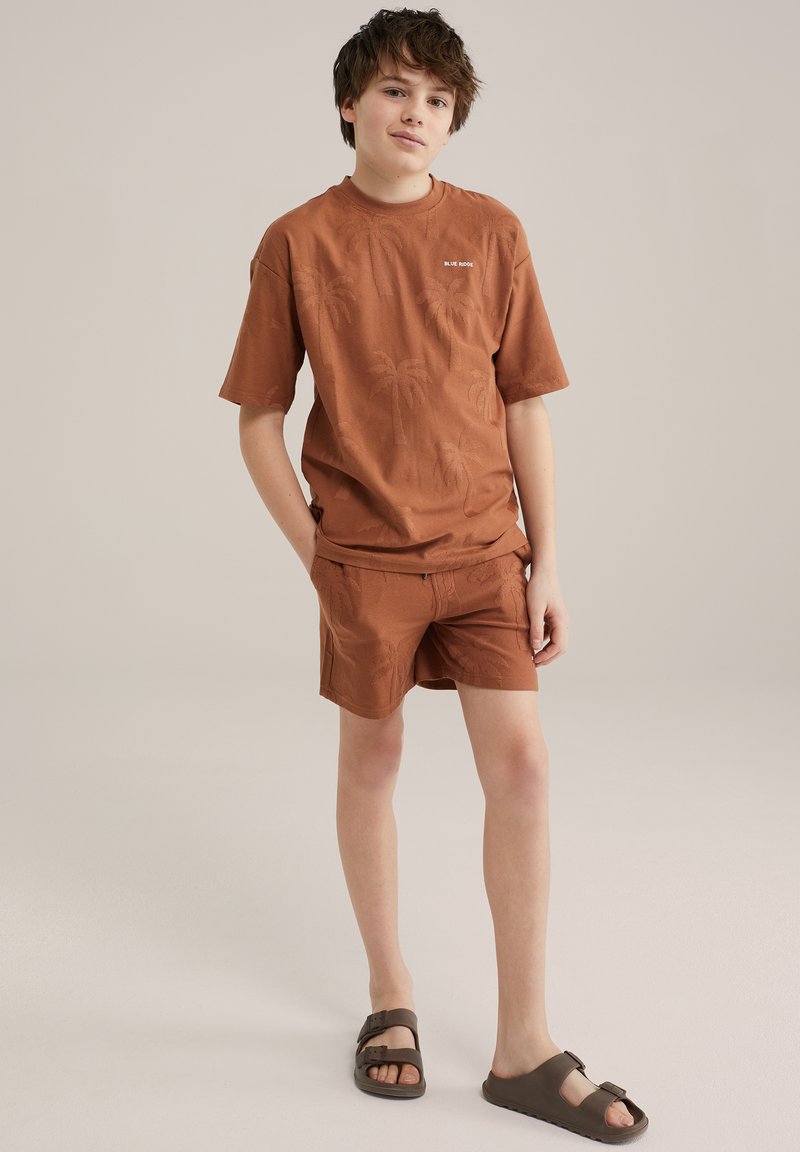 Hnedé bavlnené tričko s krátkymi rukávmi so vzorom paliem, skombinované s rovnakými šortkami. Hnedé sandále dopĺňajú celý outfit.
