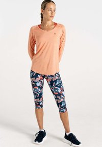 Helles orange Langarmshirt, kombiniert mit navyblauen Caprihosen mit floralem Muster. Die Schuhe sind schwarze Sneakers mit weißen Sohlen. Glatte Textur insgesamt.