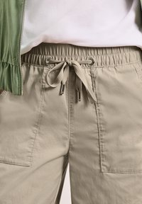 Beżowe szorty z elastycznym pasem i sznurkiem zawiązanym na kokardę, noszone z białą koszulą i zieloną kurtką na zamek.