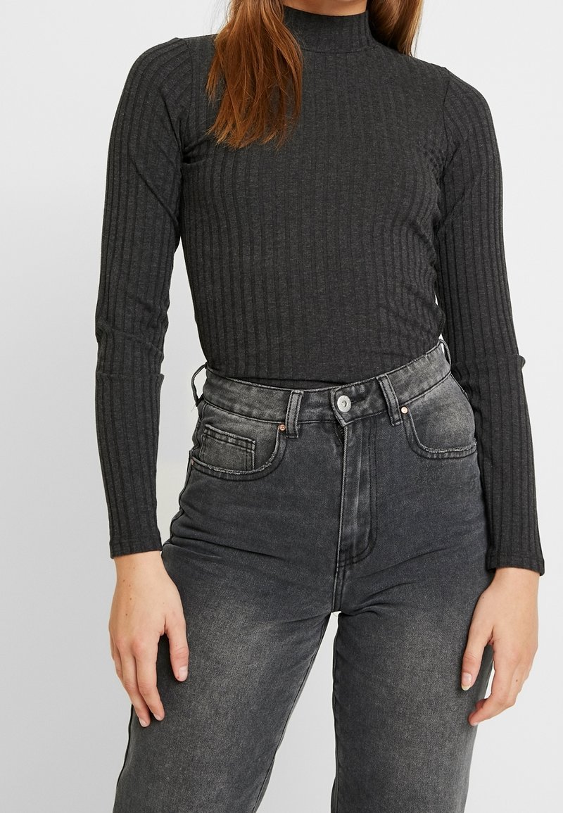 Personne portant un haut gris foncé ajusté, côtelé, à manches longues, rentré dans un jean noir délavé taille haute avec poches avant et fermeture bouton.
