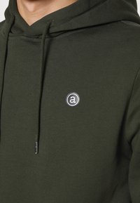 Felpa verde oliva con coulisse, caratterizzata da un logo circolare sul petto. Il tessuto ha una consistenza liscia e un design aderente.