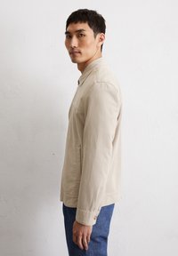 Light beige button-up jacket with a collar, side pockets, and long sleeves, made from a smooth fabric. Worn over blue denim pants.