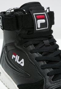 Baskets montantes noires FILA avec patchs logo blancs et rouges, lacets noirs et sangle rembourrée à la cheville sur semelle blanche.