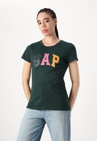 Žalias medvilninis marškinėliai su apvaliu kaklu ir trumpomis rankovėmis, su ryškiomis daugiaspalvėmis raidėmis "GAP" ant krūtinės.