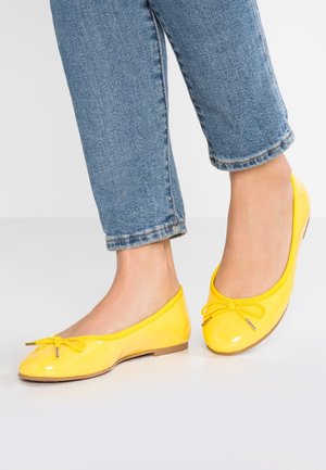 Pieds portant un jean bleu et des ballerines jaune vif avec un détail nœud sur une surface claire.