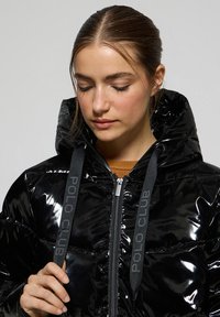 Svart puffjacka med glansig yta, hög krage och grå "POLO CLUB"-logotyp på dragsnörena. Dragkedjestängning, vadderad struktur.