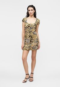 Frau steht und trägt ein kurzärmeliges, quadratisches Ausschnitt, figurbetontes Minikleid mit schwarzen, beigen und senffarbenen abstrakten Mustern und schwarze Riemchenabsätze.