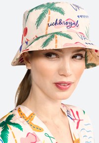 Klobouk typu bucket vyrobený z lehké tkaniny, s živým tropickým vzorem s palmami, třešněmi a nápisem v modré barvě napsaným rukou.