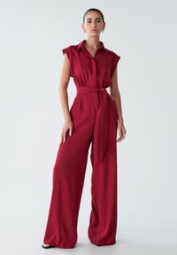 Burgundin jumpsuit, jolla on lyhyet hihat, nappisuljenta edessä ja vyöllinen vyötärö. Mukana leveät lahkeet sileässä, virtaavassa kankaassa.