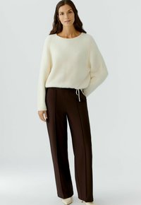 Cremfarbener Strickpullover mit runden Ausschnitt, kombiniert mit braunen, hochgeschnittenen Hosen, die seitliche Paspelierungen und einen elastischen Bund haben.