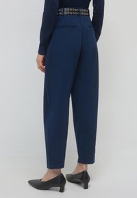 Persona che indossa pantaloni a vita alta taper blu navy con una cintura nera e scarpe nere a tacco basso, in piedi su uno sfondo neutro.