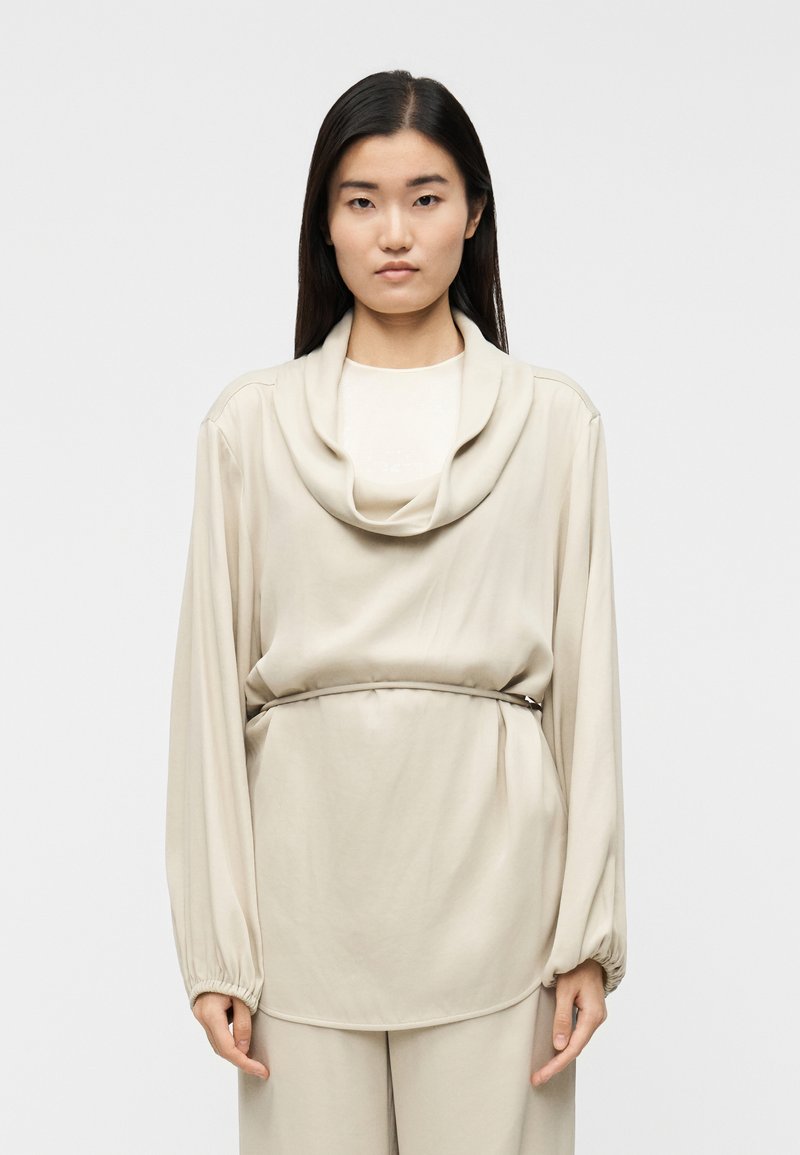 Μπεζ μπλούζα με μακρύ μανίκι και ντραπέ λαιμόκοψ η, με ελαστικά τελειώματα στα μανίκια και ελαφριά ροή. Διαθέτει κορδέλα στη μέση και ομαλή, μαλακή υφή.