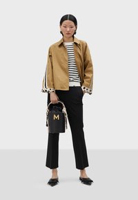 Braune Jacke mit gestreiften Ärmeln, schwarz-weißer gestreifter Pullover, schwarze Hose, schwarze Beuteltasche mit goldenen "M" und schwarze spitze Ballerinas.