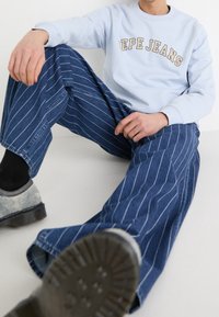 Sudadera azul claro con "Pepe Jeans" bordado en beige, combinada con pantalones vaqueros de rayas anchas azules. Zapatos grises con suelas texturizadas.