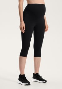 Črne nosečniške capri legice z visokim pasom, izdelane iz gladkega, elastičnega materiala, v kombinaciji s črnimi športnimi čevlji z belimi podplati.