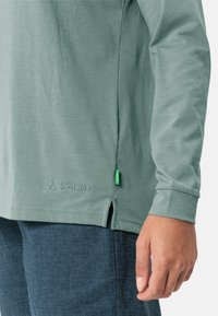 Langarmshirt aus gedämpftem grünem Stoff, mit seitlichen Schlitzen und gesticktem "VAUDE"-Logo. Besitzt einen kleinen grünen Akzent am Saum.