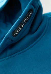 Een hoodie gemaakt van een levendig teal stof, met een zachte textuur, een trekkoordkap en een zwart label met witte tekst op de kraag.