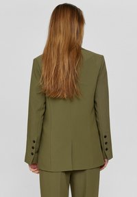 Olive roheline ülikonnajakk, millel on kohandatud lõige, ainus lõhik ja neli musta nuppu varrukaotsas. Sile kangatekstuur, sirge selja disain.