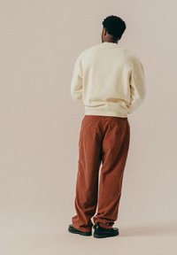 Osoba z krótkimi kręconymi włosami, ubrana w kremowy sweter, brązowo-czerwone w kratkę spodnie, czarne buty, stojąca tyłem na jednolitym beżowym tle.