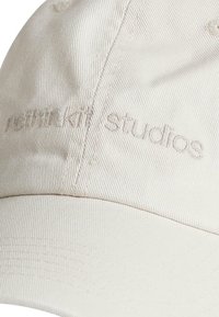 Cappello in cotone color crema con ricamo del testo "revnikat studios", dalla forma strutturata e superficie testurizzata.