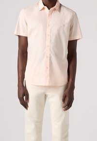 Camisa de manga corta color rosa claro con botones y cuello; presenta un pequeño parche con logo en el pecho; combinada con pantalones beige claro.