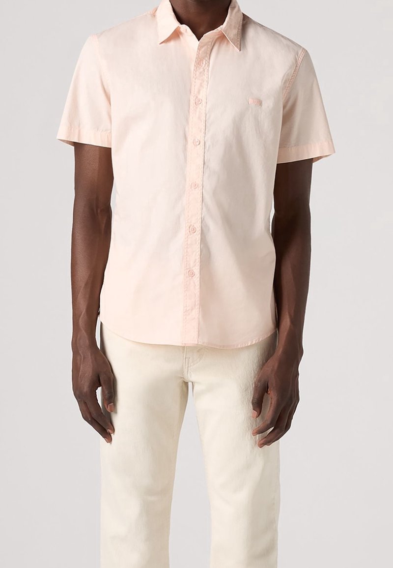 Camisa de manga corta color rosa claro con botones y cuello; presenta un pequeño parche con logo en el pecho; combinada con pantalones beige claro.