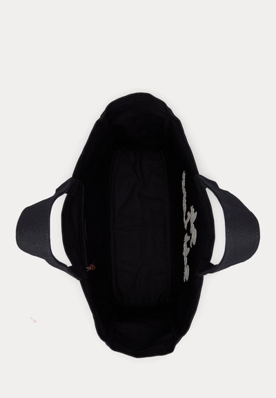 Μαύρη υφασμάτινη τσάντα tote με ευρείς λαβές, εμφανίζεται από πάνω, με άδειο εσωτερικό και μερικό λευκό κεντητό σχέδιο σε μία εσωτερική πλευρά.