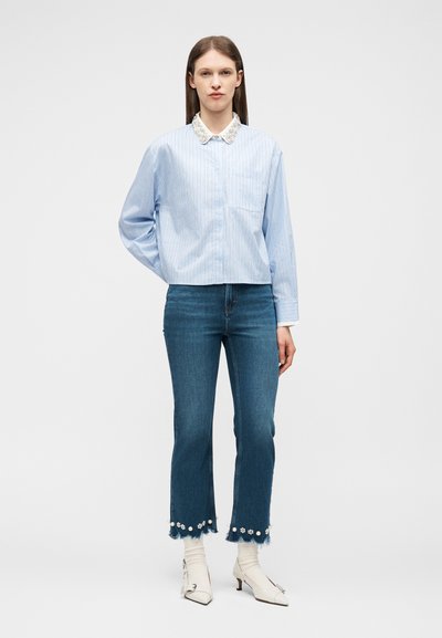 Γυναίκα που φοράει ανοιχτό μπλε πουκάμισο με ρίγες και λευκή δαντελωτή γιακά, cropped τζιν με φλοράλ λεπτομέρειες, λευκές κάλτσες και λευκά παπούτσια με τακούνι.