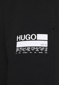 Schwarzer Stoff mit einem weißen aufgedruckten Logo, das "HUGO" liest, sowie beschreibendem Text darunter. Die Textur wirkt glatt und weich.