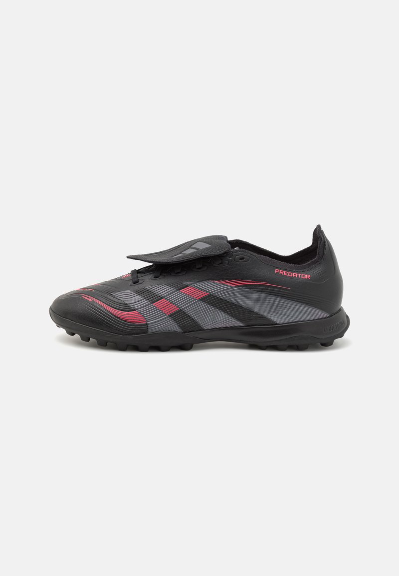 Chaussures de football synthétiques noires et grises avec des accents rouges, présentant un design élégant, une surface texturée et une sangle sur les lacets.