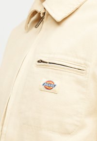 Μπεζ μπουφάν Dickies με φερμουάρ στην τσέπη στο στήθος και λεπτομέρεια στο γιακά.