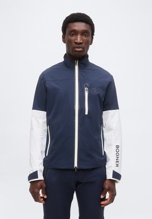 Homme portant une veste zippée bleu marine et blanche avec "BOGNER" sur la manche gauche, debout devant un fond clair uni.