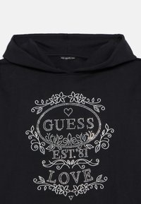 Svart hettegenser med sølvfarvede rhinestone-pynt. Designet inkluderer blomster mønstre og teksten "GUESS EST. 81 LOVE" i stiliserte skrifttyper.