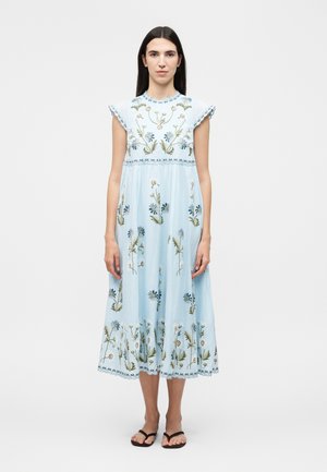 Femme portant une robe midi bleu clair avec une broderie florale et des manches courtes, debout pieds nus devant un fond uni.