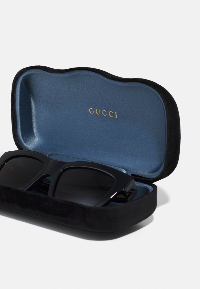 Lunettes de soleil noires dans un étui en velours noir, avec un intérieur bleu arborant le logo doré "GUCCI". L'étui a un design à bord ondulé.