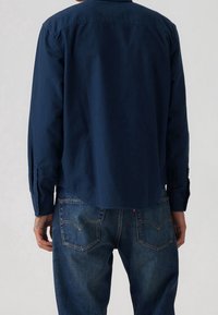 Granatowa koszula na guziki wykonana z bawełny, z długimi rękawami zakończonymi mankietami na guziki, prostym dołem i miękką fakturą. Noszona z ciemnymi jeansami.