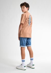 Camiseta de algodón color marrón claro con estampado gráfico azul y naranja en la parte posterior, combinada con pantalones cortos de mezclilla enrollados y zapatillas blancas con acentos verdes.