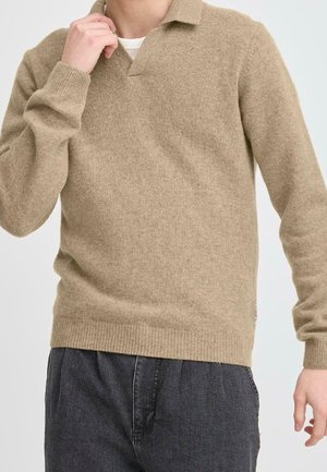 Person iført en beige strikket sweater med krave og mørkegrå bukser, den ene hånd nær kraven og den anden ved siden.