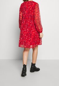 Rotes Blumenkleid mit durchsichtigen langen Ärmeln, tailliertem Schnitt und knielangem Saum, kombiniert mit schwarzen Knöchelstiefeln vor einem hellen Hintergrund.
