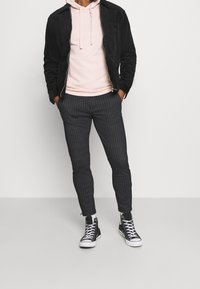Man wearing black zipped jacket over light pink hoodie, dark gray pinstripe pants, white socks, and black high-top sneakers standing with hands in pockets.