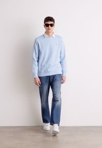 Light blue sweatshirt with a white collar, paired with faded blue jeans and white sneakers. Model wears dark sunglasses; plain background.