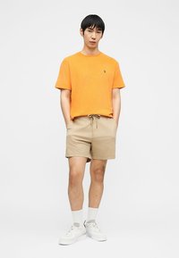 Ung mann som står med hendene i lommene iført oransje kortermet skjorte, beige shorts med snor, hvite sokker og hvite joggesko.