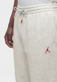 Ανοιχτόγκρι φόρμα με κόκκινο λογότυπο Air Jordan κοντά στην τσέπη και λευκό κορδόνι με κόκκινα άκρα δεμένο σε φιόγκο σε άτομο με σκουρόχρωμο δέρμα.