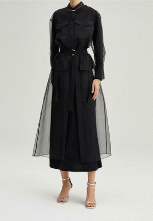 Vestito nero a balze con sovrapposizione trasparente, chiusura frontale con bottoni, tasche sul petto e cintura in vita. Il look è completato da sandali argentati con cinturini.