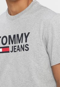 Gray cotton t-shirt with a crew neckline, featuring prominent black text "TOMMY" above “JEANS” and a horizontal red and white stripe design.